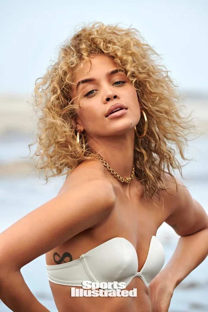 Jasmine Sanders poses in front of the ocean in a white strapless bikini top and gold necklace.